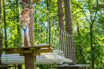 A little girl wearing a safety harness, standing on a wooden platform in an adventure park, holding...
