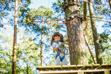 Cute little girl standing on a wooden platform at a high ropes course, holding ropes and carabiners, in an adventure park