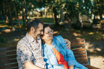 Elderly pair seated on a bench under the shade of trees, embracing the calmness of the natural...