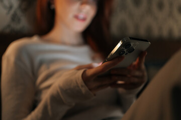 Fototapeta na wymiar Close-up cropped shot of woman relaxing in dark bedroom, scrolling social media on smartphone, screen warm glow illuminating face. Calm female browsing Internet using mobile phone looking to screen.