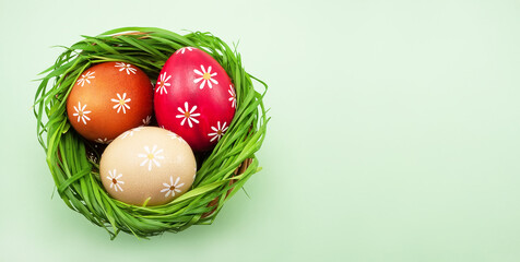 Colored Easter eggs in a basket with green grass on a light green colored table, top view
