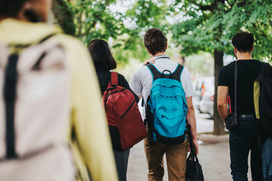A group of friends wearing backpacks walk together on a tree-lined sidewalk, enjoying a sunny day outdoors. Captures a sense of camaraderie and adventure. - Powered by Adobe