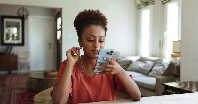 Serious beautiful young African woman leaving voice message on smartphone, keeping mobile phone at mouth, speaking, getting happy, smiling, finishing talk, typing, sitting at home table