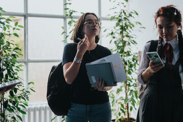 Two students are conversing and using a smart phone in a bright hallway filled with plants. The...