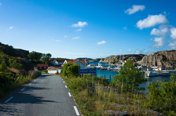 Fototapeta premium Scenic coastal road in Hamburgsund, Sweden, with a view of the marina, sailboats, and red-roofed houses. A peaceful summer landscape with rocky cliffs and lush greenery under a blue sky