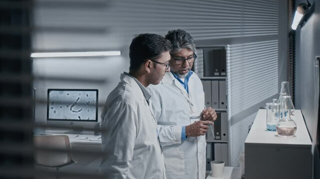 Side shot of mature Indian scientist talking to young male assistant, teaching him to work with solutions and reagents in modern laboratory
