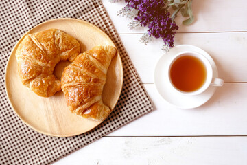 Cup of tea with purple flower and wooden dish with croissants on white wooden background.