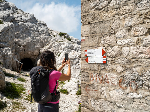 A hiker takes a photo of a trail marker on a stone wall. The marker has red and white arrows pointing to different destinations. A mountain and a dark cave entrance are in the background. Flower path,
