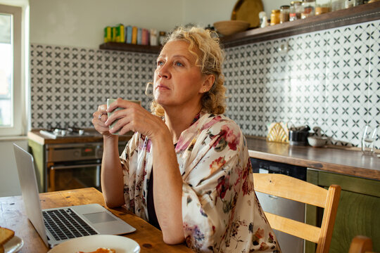 Middle aged woman drinking coffee and looking thoughtful in a cozy kitchen - Powered by Adobe