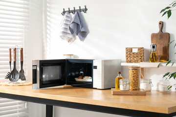 Open microwave oven with food and kitchen utensils on wooden table indoors
