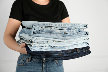 Woman with stack of different stylish jeans near light wall indoors, closeup