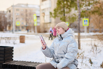 Fototapeta premium Portrait young Caucasian girl using smartphone, looking at mobile phone screen on winter walk in park