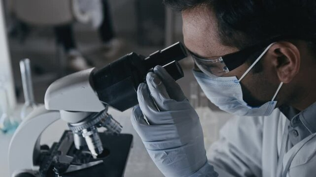 Side close-up slowmo shot of face of male lab assistant in protective goggles, face mask and gloves studying sample under microscope while making experiments in drug development laboratory