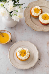Lemon Poppy Seed Cupcakes with lemon curd and cream cheese topping. Cupcake in form of Fried Egg. Colourful Easter eggs, flowers on a stone background, top view. Easter holiday and festive food.