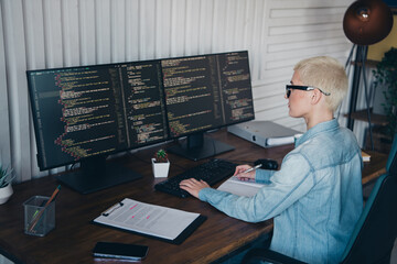 Focused female programmer working on dual monitors coding and developing software in a modern home...