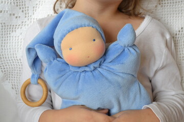 Child holding blue Waldorf doll with wooden ring and joyful expression on white background
