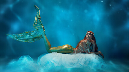 Mystical mermaid lying on sheer floating fabric, her golden tail floating, she gazing dreamily into...