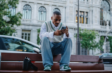 young Black man in a fashionable outfit sits on a wooden bench, engaging with his smartphone and...