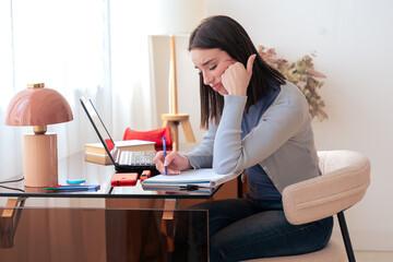 Caucasian student studying at home, writing in notebook while using laptop. Modern workspace with stylish decor, lamp, and stationery. Education, learning, productivity, and concentration.