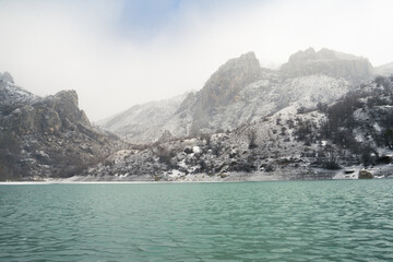 Obraz premium Lake in the snow-covered mountains with a deep turquoise hue in misty clouds. Winter landscape. Magic of wild nature. Snowflakes gently fall. Travel, adventure, journey, ecotourism, meditation, hiking