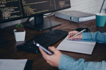 A young professional writing notes by a computer screen while multitasking in a modern workspace