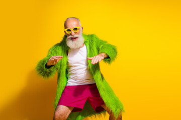 Stylish senior man in vibrant green fur jacket and yellow sunglasses posing against yellow...
