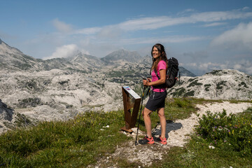 A woman with a backpack and hiking poles stands on a mountain trail, smiling. Mountains and a partly cloudy sky are in the background. Flower path, Julian natural Park, Gilberti hut, Sella Nevea.