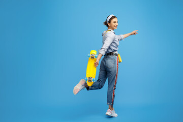 Energic young woman with yellow skateboard dancing on blue background