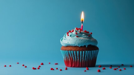 Blue cupcake with red sprinkles and lit candle on blue backdrop.