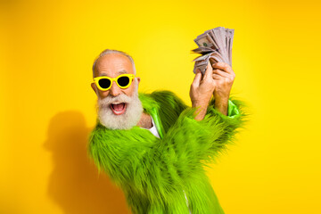 Charismatic elderly man wearing vibrant fur and sunglasses celebrating wealth on yellow background
