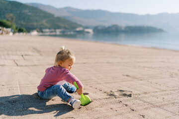 Little girl sits on a sandy beach and digs the sand with a toy shovel