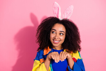 Cheerful woman wearing bunny ears poses with a playful gesture against a pink background,...