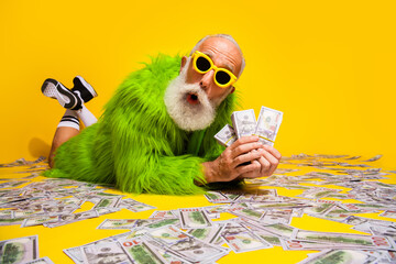 Eccentric senior man in a green fur coat posing on yellow background holding US dollars, showcasing...