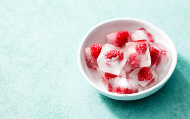 Frozen ice cubes with fresh raspberries in a white bowl. Blue background. Close up. Copy space.