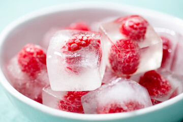 Frozen ice cubes with fresh raspberries in a white bowl. Blue background. Close up.