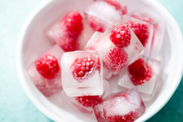 Frozen ice cubes with fresh raspberries in a white bowl. Blue background. Close up.