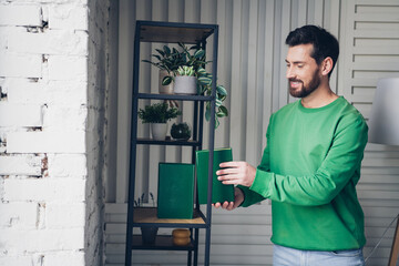 Smiling man organizing green books on metal shelf in stylish indoor space