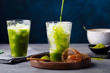 Matcha, green tea ice tea pouring in tall glass on wooden plate. Grey and blue stone background. Close up.