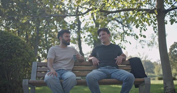 Two friends enjoy a relaxing afternoon in the park, chatting and drinking coffee on a bench under the shade of a tree.