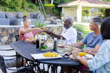 Senior Friends enjoying outdoor meal, sharing food and conversation in sunny backyard