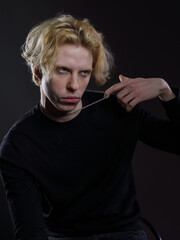 Portrait of a young man with wavy blonde hair in black clothing against a dark background. His expression is serious, and his gaze is intense. Dramatic lighting highlights his facial features and hair