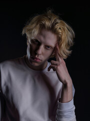Portrait of a young man with wavy blonde hair in white clothing against a dark background. His expression is serious, and his gaze is intense. Atmospheric lighting highlights facial features and hair 