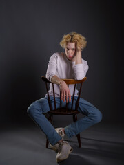 Portrait of a young man with wavy blonde hair in white clothing against a dark background. His expression is serious, and his gaze is intense. Atmospheric lighting highlights facial features and hair 