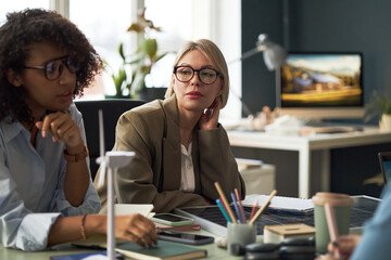 Young woman sitting at table at meeting and listening to her colleagues
