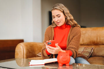 Young woman managing finances while using smartphone and sitting on a couch in a cozy living room...