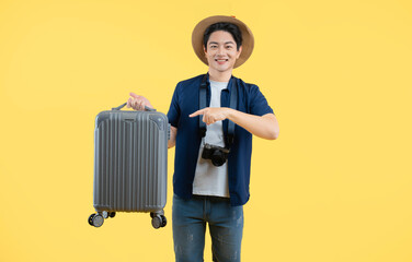Portrait of a young Asian man wearing a camera and carrying a travel suitcase posing on a yellow background