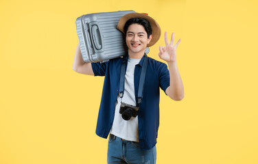 Portrait of a young Asian man wearing a camera and carrying a travel suitcase posing on a yellow...