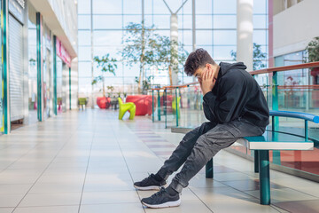 Fototapeta premium eenager in black hoodie sitting on a bench with head in hands, feeling stressed or overwhelmed, sensory overload, autism, ADHD, bullying, mental health in a bright shopping mall. Copy space