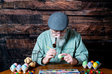 old man with grey beard smoking pipe painting eggs for Easter with dark wood background