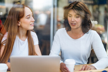 Two women having productive conversation while working on laptop in busy urban cafe during the afternoon